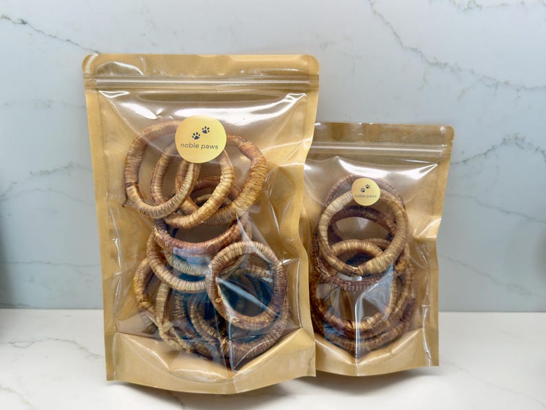 May include: Two clear plastic bags filled with dog treats. The bags are brown and sealed with a zipper. The treats are ring-shaped and brown. Each bag has a gold sticker with the words "noble paws" and paw prints.