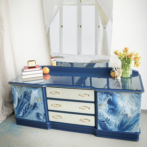 Puede incluir: Un tocador azul con un diseño de mármol blanco y detalles dorados. El tocador tiene tres cajones y una puerta de armario. Tiene una tapa de vidrio y un espejo grande y curvo encima. Hay algunos libros, una naranja y un jarrón de flores en la parte superior del tocador.