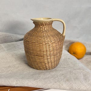 Puede incluir: Un jarrón de mimbre tejido con un acabado marrón claro y una parte superior de cerámica crema. El jarrón tiene un asa y un cuerpo redondeado.