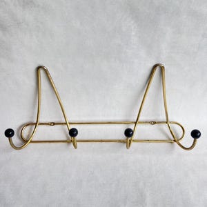 Puede incluir: Un perchero de pared vintage de latón con tres ganchos negros en forma de bola. El perchero tiene un diseño decorativo con dos triángulos que apuntan hacia arriba y una barra horizontal.
