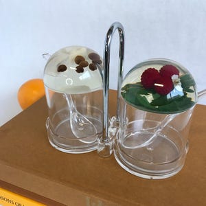 May include: A clear acrylic condiment holder with two glass containers. The containers have lids with a dome shape. One container has coffee beans inside and the other has a red raspberry and green leaves. The holder has a chrome metal handle.