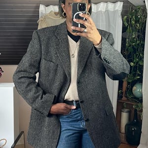 May include: A gray tweed blazer with black elbow patches, paired with blue jeans. The blazer has a classic design with a notched lapel and buttoned cuffs. The outfit is completed with a black belt and a light-colored sweater.