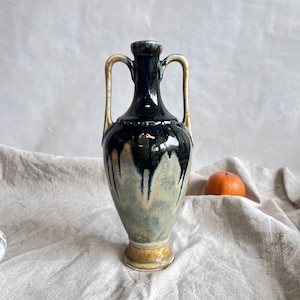 May include: A small, blue and white ceramic vase with two handles. The vase has a drip glaze effect and a rounded body.