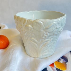 May include: White ceramic planter with a floral design. The planter is empty and sitting on a white fabric surface.