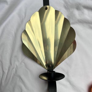 Könnte beinhalten: Ein Wandkerzenhalter mit einem goldfarbenen, fächerförmigen Design. Der Kerzenhalter hat einen schwarzen Metallsockel und eine Schlaufe zum Aufhängen. Die Fächerform besteht aus einem glänzenden, metallischen Material. Das Gesamtdesign ist vintage und dekorativ.