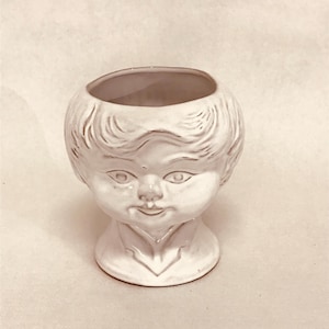 May include: White ceramic planter shaped like a person's head with a smiling face. The planter has a round top and a base.