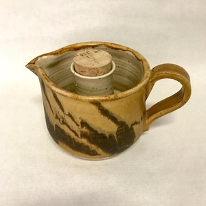 May include: A handmade ceramic creamer with a cork stopper. The creamer is tan with dark brown streaks and a curved handle. The cork stopper is in a cylindrical opening in the center of the creamer.