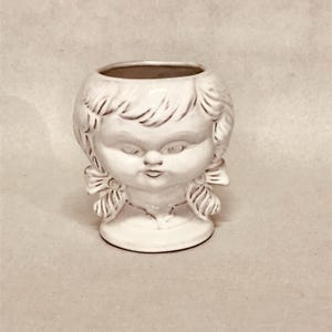 May include: White ceramic planter shaped like a girl's head with pigtails. The planter has a simple design and a smooth, matte finish.