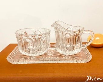 May include: A clear glass sugar bowl and creamer set with a decorative, cut-glass pattern. The set is on a clear glass tray with a similar pattern. The tray is sitting on a brown book.