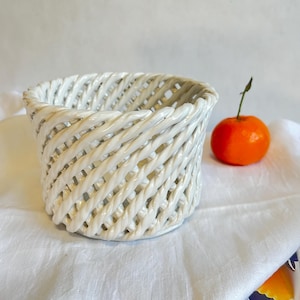 May include: White ceramic basket with a woven design. The basket is empty and sitting on a white fabric surface.