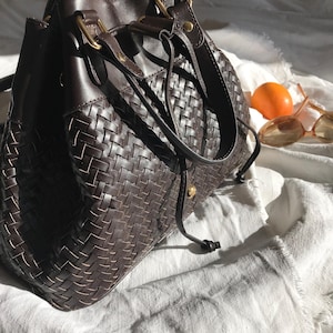 May include: A brown woven leather bucket bag with gold hardware and a drawstring closure. The bag is sitting on a white fabric surface.