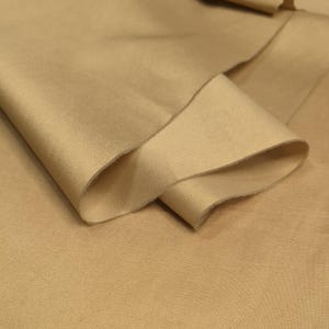 May include: Close-up of a piece of tan-colored fabric with a textured surface. The fabric is folded over, revealing a raw edge and the underside of the material. The lighting highlights the subtle variations in the fabric's color and texture.