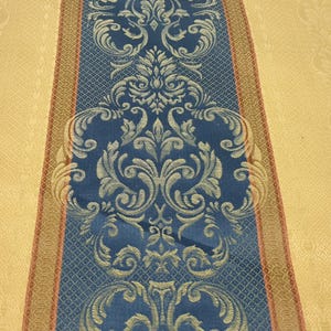 May include: A close-up of a decorative textile with a central blue panel featuring a detailed, ornate floral design in silver and gold. The blue panel is bordered by a gold and red stripe, set against a yellow background.