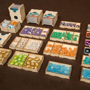 Könnte beinhalten: Ein Satz aus Holzaufbewahrungsboxen zum Organisieren von Spielsteinen. Die Boxen sind aus hellem braunem Holz gefertigt und haben verschiedene Fächer für die Aufbewahrung von Spielsteinen unterschiedlicher Größe. Die Boxen sind mit bunten Spielsteinen gefüllt, darunter grüne, blaue, orange und weiße.
