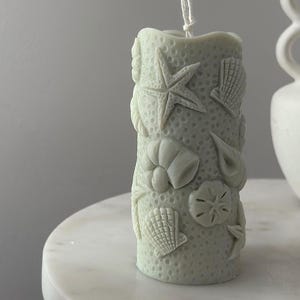 May include: A light green cylindrical candle with seashell and starfish designs. The candle has a textured surface and a white wick. The candle is on a white marble table.
