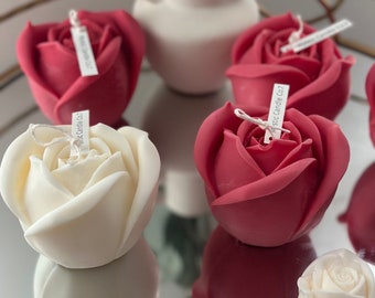 May include: Five rose-shaped candles, three in a deep pink shade and two in white, are arranged on a mirrored surface with a gold trim. The candles are all in the shape of a rose bud, with a small tag attached to each one.