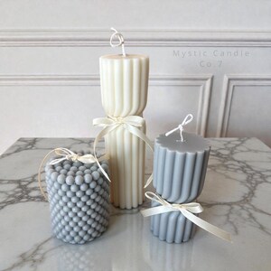 May include: Four decorative candles in varying shapes and colors. The candles include a tall, ribbed ivory candle, a gray candle with a bubble pattern, and two gray ribbed candles. Each candle is tied with a white ribbon. The text "Mystic Candle Co. 7" is visible.