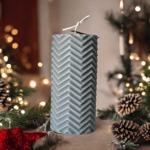 May include: A gray pillar candle with a chevron pattern. The candle is sitting on a wooden surface with pine branches and pine cones.