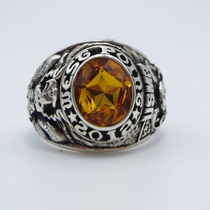 Peut inclure: Une bague en argent avec une grande pierre précieuse facetée de couleur ambre. La bague présente un motif d'aigle et l'inscription "POLICE EMS 1926".