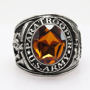 Peut inclure: Une bague en argent avec une grande pierre précieuse orange au centre. La bague est gravée des mots "U.S. ARMY PARATROOPER".