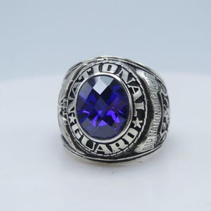 Peut inclure: Bague argentée ornée d'une pierre précieuse ovale, facettée, bleu foncé au centre. La bague est gravée des mots "AIR NATIONAL GUARD" et d'autres détails. La bague a un design texturé et détaillé.