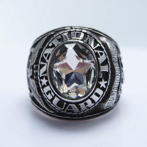May include: A silver-toned ring with the words "NATIONAL GUARD" engraved around a large, clear, star-shaped stone. The ring has intricate detailing and is set against a white background.