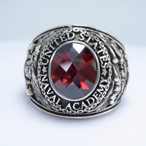 May include: A silver ring with a red gemstone and the inscription "UNITED STATES NAVAL ACADEMY".