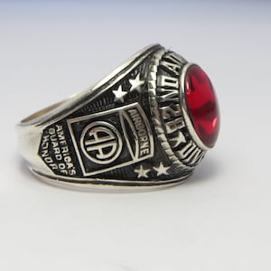Peut inclure: Une bague en argent avec une pierre rouge et l'inscription "America's Guard of Honor 258th Airborne".