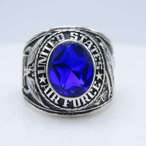 May include: A silver-toned ring with a large, oval, deep blue gemstone at its center. The ring is engraved with the words "UNITED STATES AIR FORCE" around the gemstone. The ring has intricate side detailing.