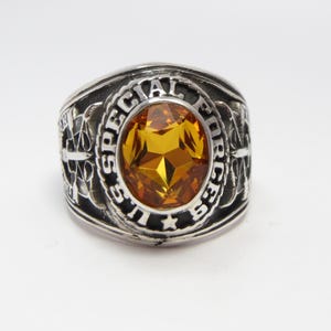 May include: A silver ring with a large amber-colored gemstone. The ring features the words "U.S. SPECIAL FORCES" around the gemstone.