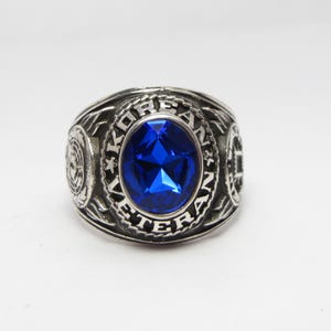 May include: A silver ring with a blue gemstone and the inscription "KOREA VETERAN" around the stone.