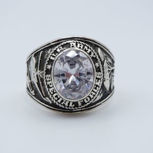 May include: A silver ring with a large clear stone in the center. The ring has the words "U.S. ARMY SPECIAL FORCES" engraved around the stone.