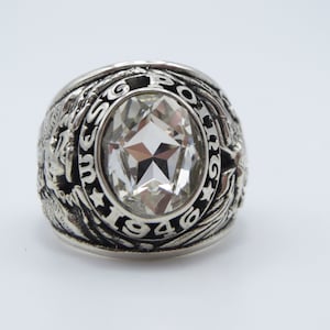 Peut inclure: Une bague argentée avec une grosse pierre transparente de taille ovale au centre. La bande de la bague présente des détails complexes, notamment du texte et des symboles. L'année "1946" est visible sur la bague.