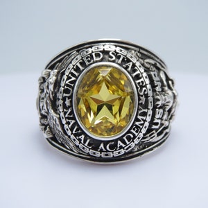 May include: A silver ring with a yellow gemstone and the inscription "UNITED STATES NAVAL ACADEMY" around the band.