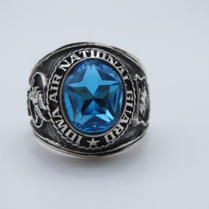 Peut inclure: Une bague argentée ornée d'une grande pierre bleue facettée en forme d'étoile au centre. L'anneau en métal est gravé du texte IOWA AIR NATIONAL GUARD, entouré d'une bordure en forme de corde et d'emblèmes en relief détaillés sur les côtés.