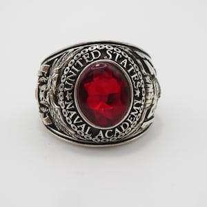 May include: A silver ring with a red stone and the inscription "UNITED STATES NAVAL ACADEMY".