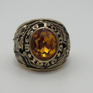 Peut inclure: Une bague de classe dorée avec une grosse pierre ambrée facetée. La bague porte l'inscription "US.S. POLARIS 1946" autour de la pierre.