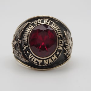 May include: A gold ring with a red gemstone and the inscription "TRUONG VO BI QUOC GIA VIET NAM" around the band.
