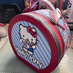 May include: A red and blue Hello Kitty themed round box with a handle. The box has a white and blue striped pattern with Hello Kitty holding a camera. The text "Hello Kitty" is printed on the box.