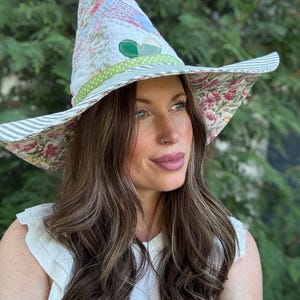 May include: A pointed hat constructed from quilted fabric featuring a patchwork of floral and patterned textiles in soft pastel colors. The hat includes a green polka-dot band and a wide brim with a striped and floral underside design.