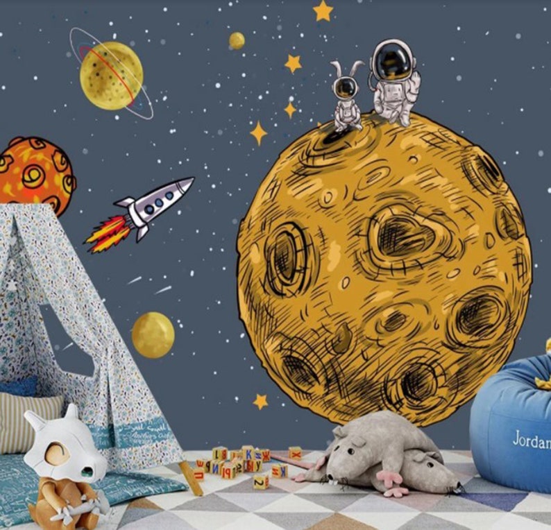 K&ouml;nnte beinhalten: Ein blaues und gelbes Wandbild zum Thema Weltraum mit einer Rakete, Planeten, Sternen und zwei Astronauten, die auf einem gro&szlig;en Mond stehen. Das Wandbild befindet sich in einem Raum mit einem Tipi, einem Stofftier und einem Sitzsack.