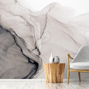 May include: Abstract wall mural with a grey and white marble-like design. The mural is displayed in a living room setting with a white armchair and a wooden side table.
