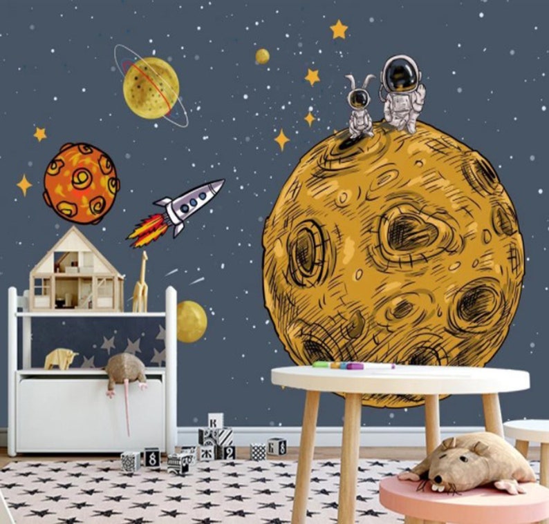 K&ouml;nnte beinhalten: Ein blaues und wei&szlig;es Wandbild zum Thema Weltraum mit einem gelben Mond, einer Rakete und zwei Astronauten auf dem Mond. Das Wandbild befindet sich in einem Kinderzimmer mit einem Holzregal, einem wei&szlig;en Tisch und einem rosa Hocker.