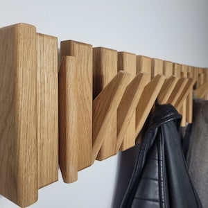 Könnte beinhalten: Ein hölzerner, an der Wand befestigter Kleiderständer mit mehreren Klapphaken. Der Ständer ist aus hellfarbenem Holz gefertigt und hat ein modernes Design. Eine schwarze Lederjacke und ein grauer Mantel hängen an den Haken.