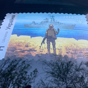 Puede incluir: Un sello postal ucraniano con un soldado de pie en una playa con un barco en el fondo. El sello es azul, amarillo y blanco con el texto "Ukraina" y "2022" impreso en él.