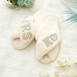 Op de afbeelding: Crèmekleurige, pluizige slippers met het woord "MRS" uitgespeld in strass op één bandje en de initialen "K♡" op de andere. De slippers liggen op een wit, getextureerd oppervlak, met bloemaccenten.