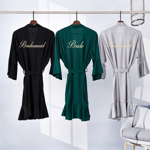 May include: Three satin robes hanging on hangers. The robe in the middle is green and says "Bride" in gold lettering. The robes on either side are black and gray and say "Bridesmaid" in gold lettering.