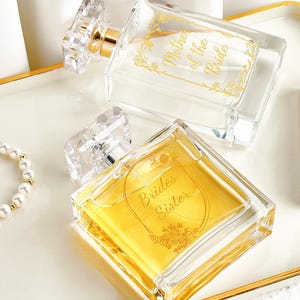 May include: A white tray with gold edging displays two perfume bottles and a lipstick. One perfume bottle is labelled "Mother of the Bride" and the other "Brides Sister". A pearl bracelet and a lipstick are also on the tray.