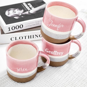 May include: Three pink ceramic mugs with white, grey, and brown bands, stacked on top of each other. Each mug has a name printed on it: "Jennifer", "Geoffrey", and "Seren". The mugs are filled with a light-coloured liquid.