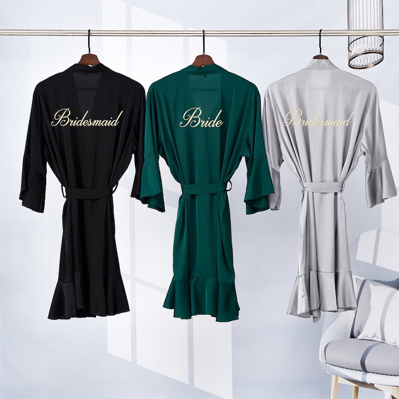May include: Three satin robes hanging on hangers. The robe in the middle is green and says "Bride" in gold lettering. The robes on either side are black and gray and say "Bridesmaid" in gold lettering.